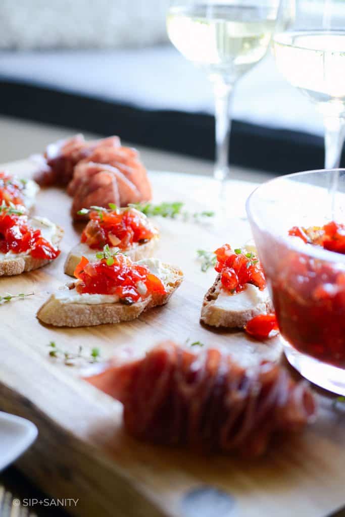 wooden board with red pepper jam crostini and white wine glasses