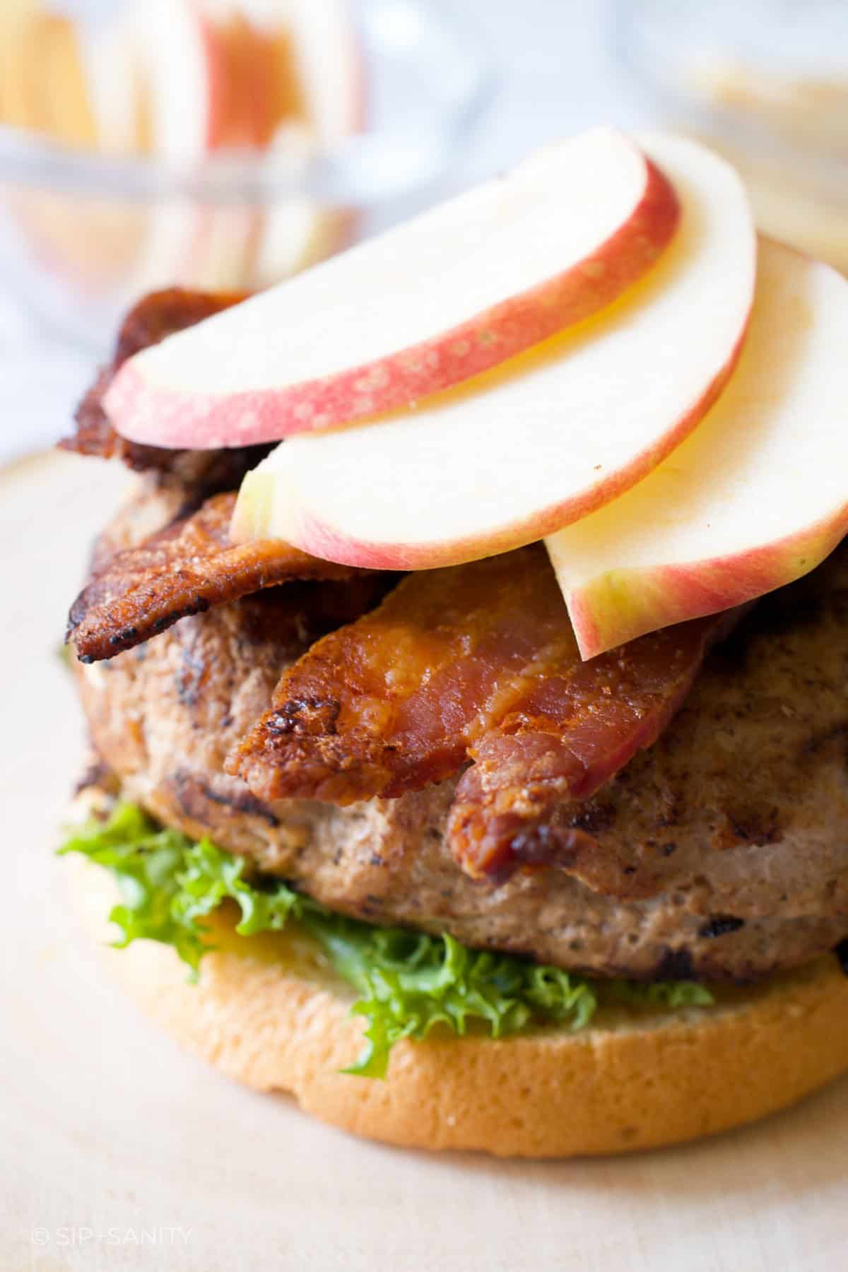 A bun, lettuce, turkey burger, bacon and apple slices stacked.