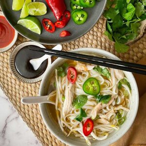 A bowl of rotisserie chicken pho next to toppings and condiments.