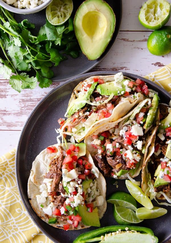 Four beef tacos on a plate next to avocado and cilantro.