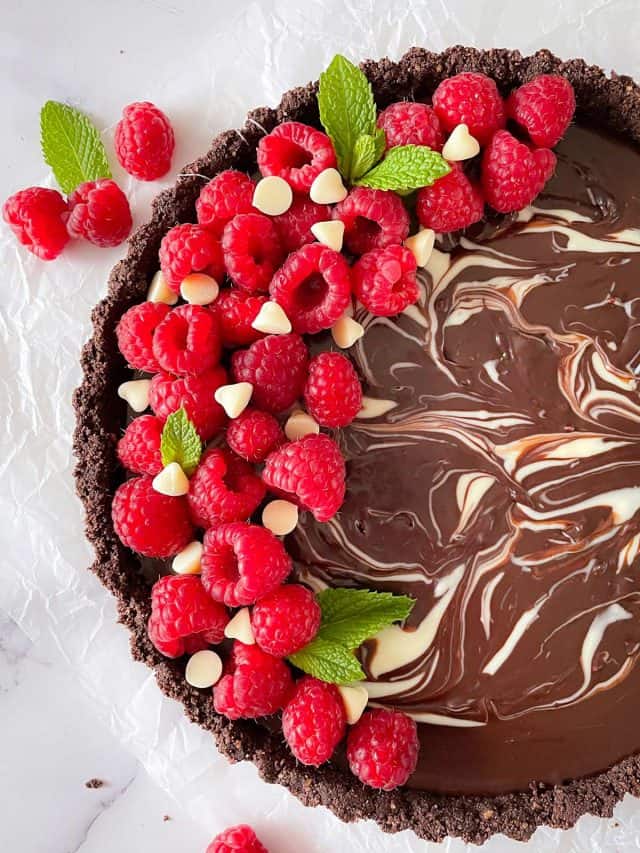 A chocolate tart with white chocolate swirls, topped with raspberries on a white countertop.