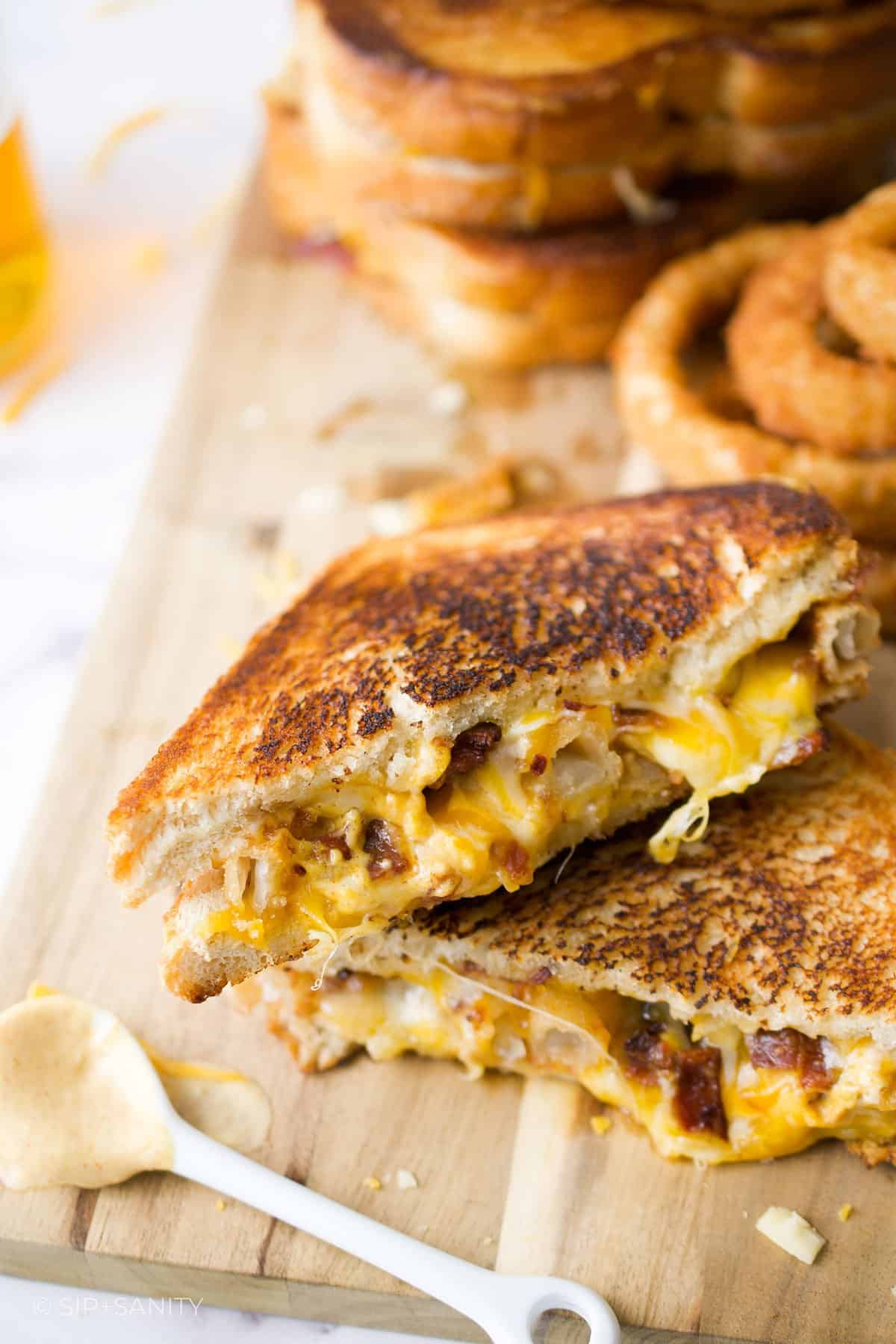 Two halves of an onion ring and bacon grilled cheese sandwich stacked on each other with more sandwiches in the background.