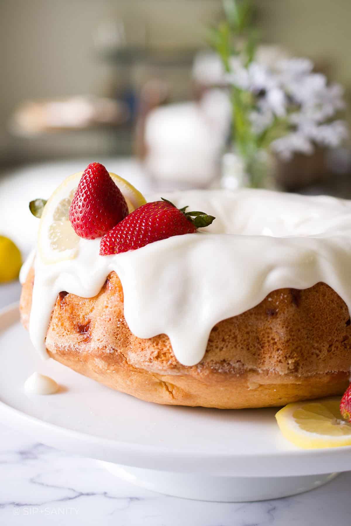A bundt cake on a platter with white icing and lemons and strawberries on top.