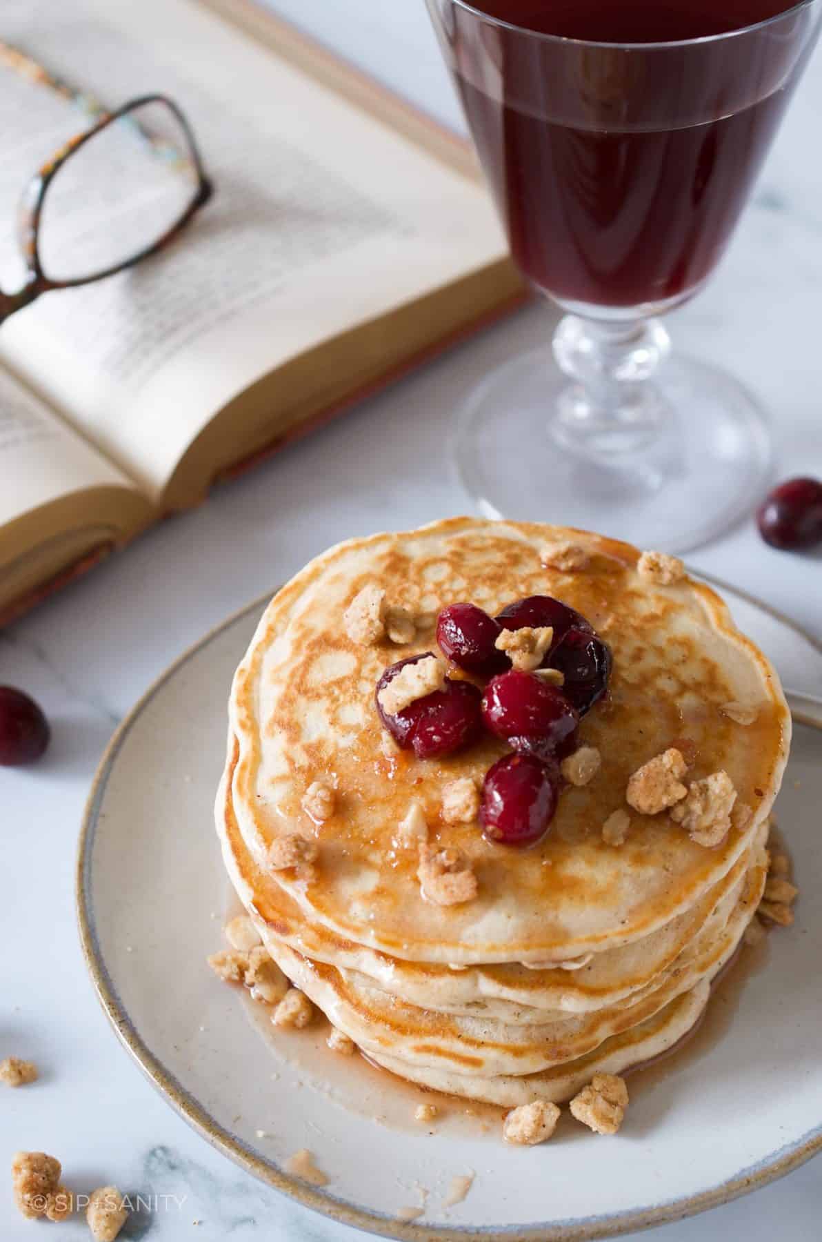 Stack of cranberry crumble pancakes next to a glass of juice and an open book.
