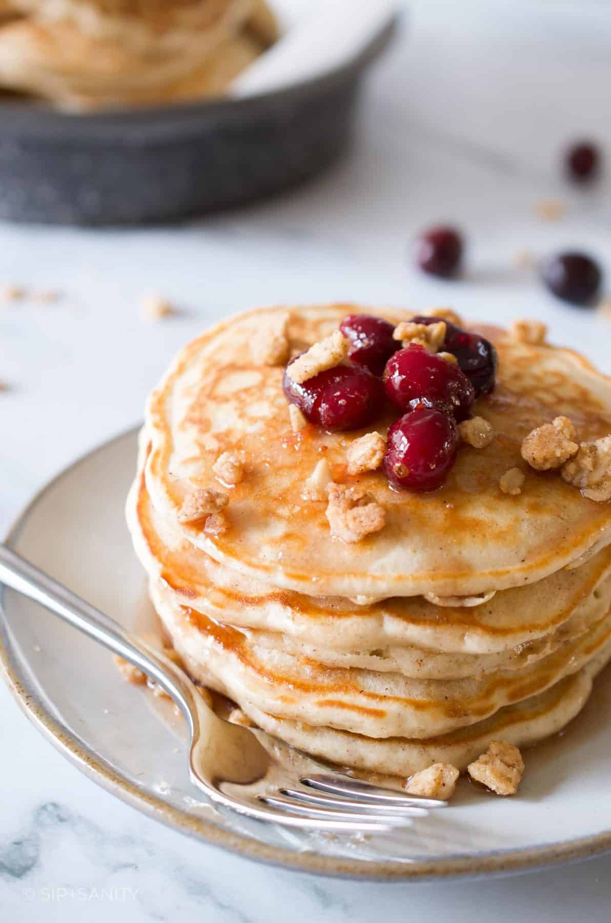 Stack of pancakes on a plate topped with cranberry maple syrup and oat crumble next to a fork.