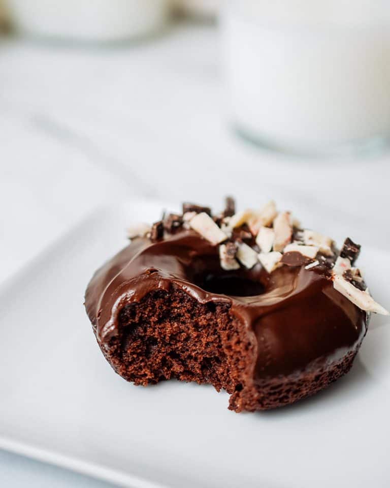 A chocolate ganache glazed chocolate donut with a bite taken from it on a white plate.