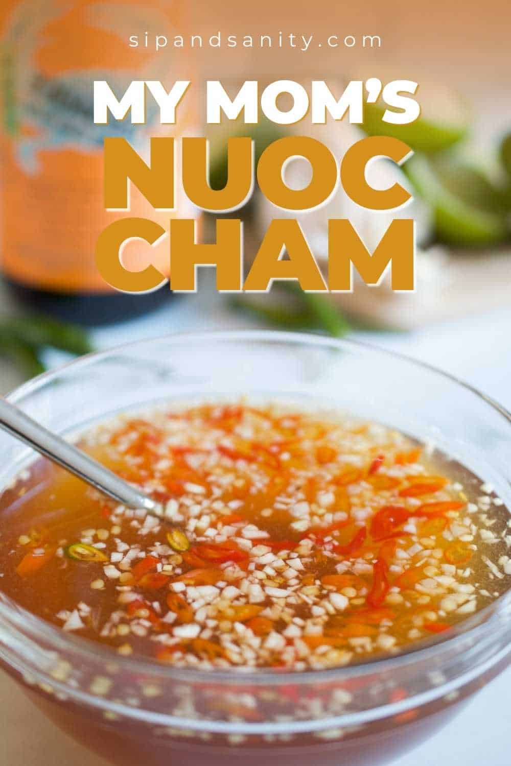 Glass bowl of Vietnamese Nước chắm with garlic, red chili, spoon. Bottle, lime wedges in back. Text: My Mom’s Nuoc Cham.