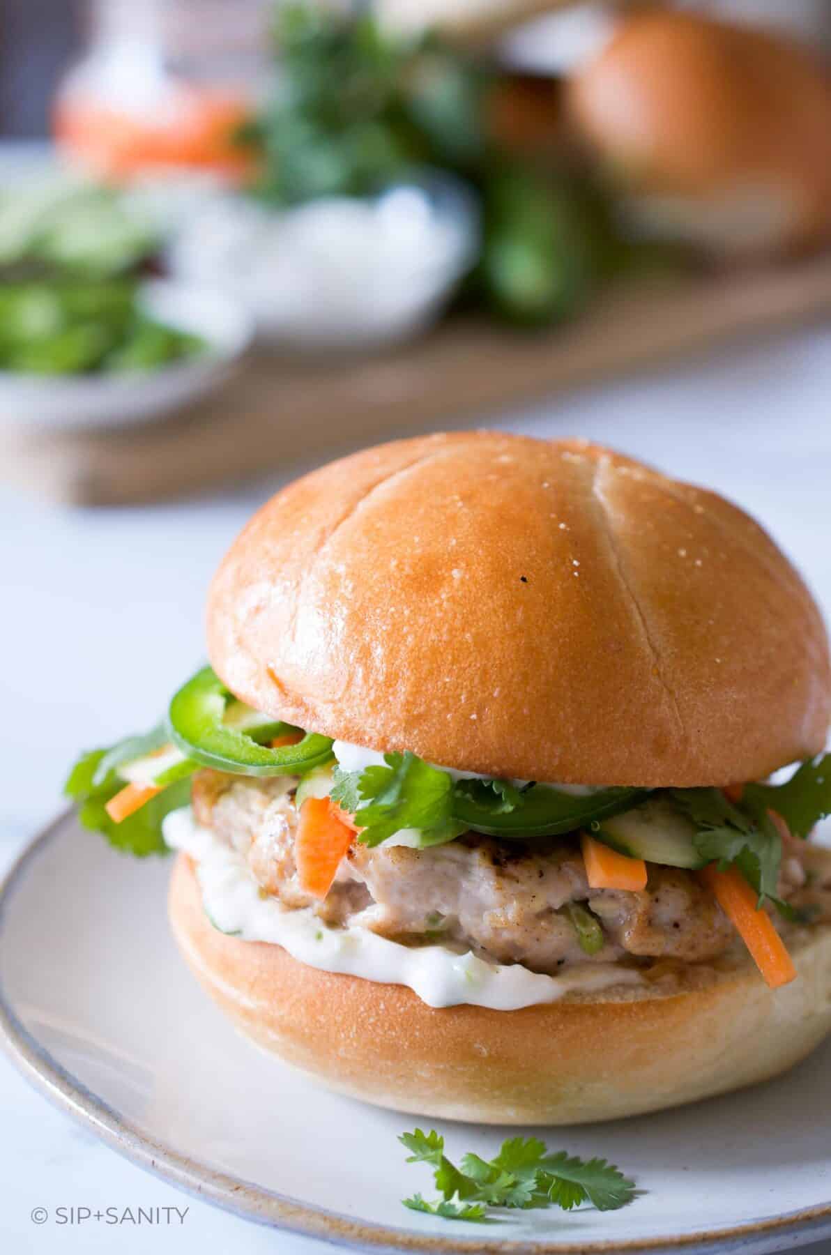 A banh mi chicken burger with scallion mayo next to a board with fresh veggies.