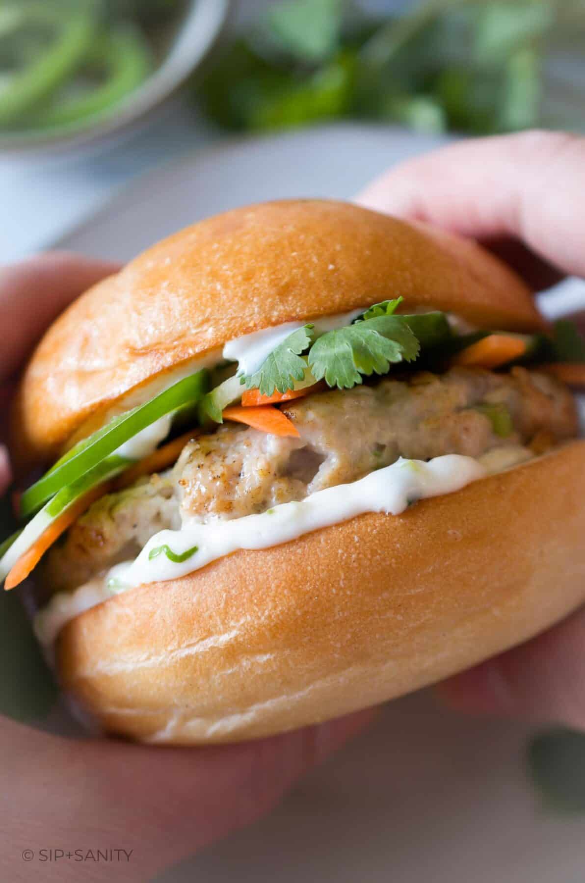 Two hands holding a banh mi chicken burger with scallion mayo.