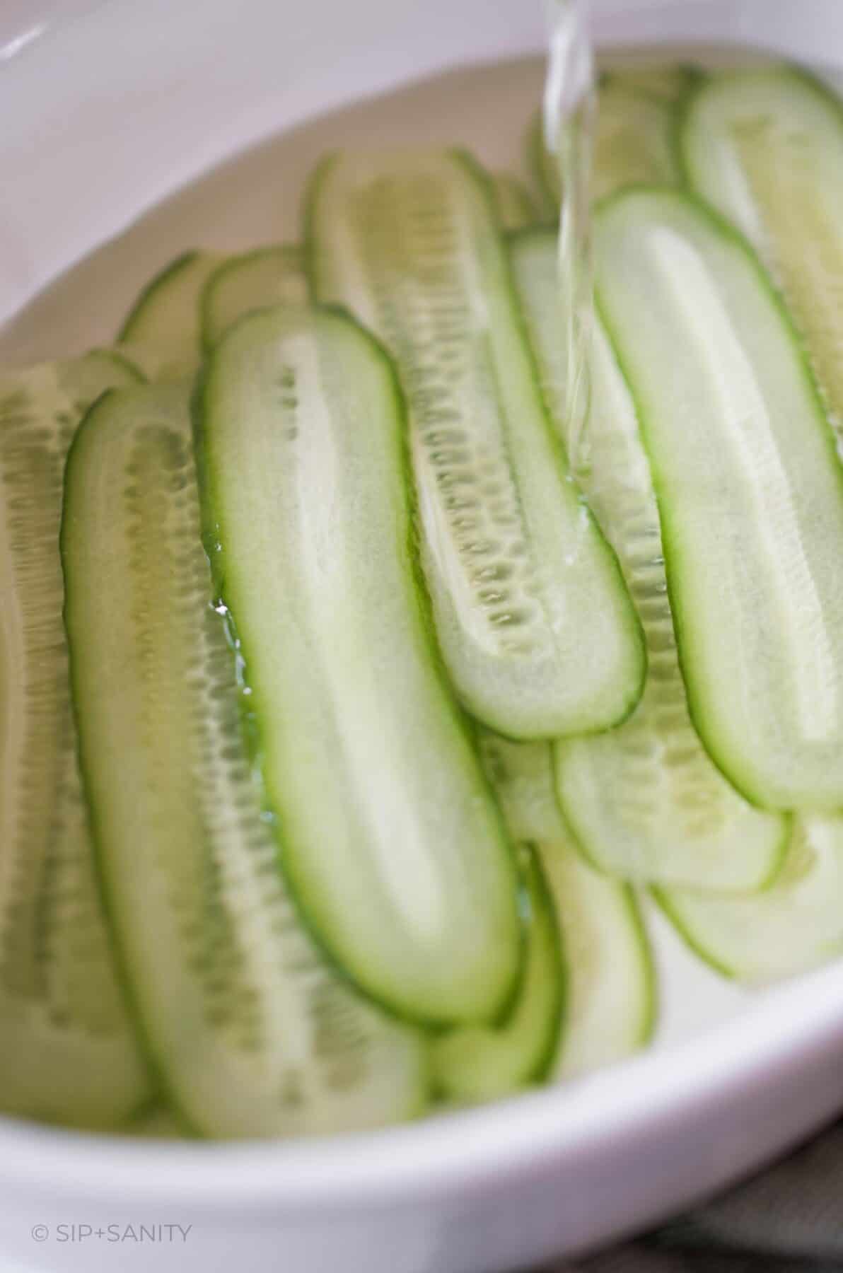 Thin slices of cucumber in a shallow dish with pickling liquid being poured over.