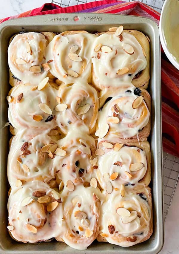 A metal baking pan filled with iced cherry sweet rolls with toasted almonds on red striped towel.