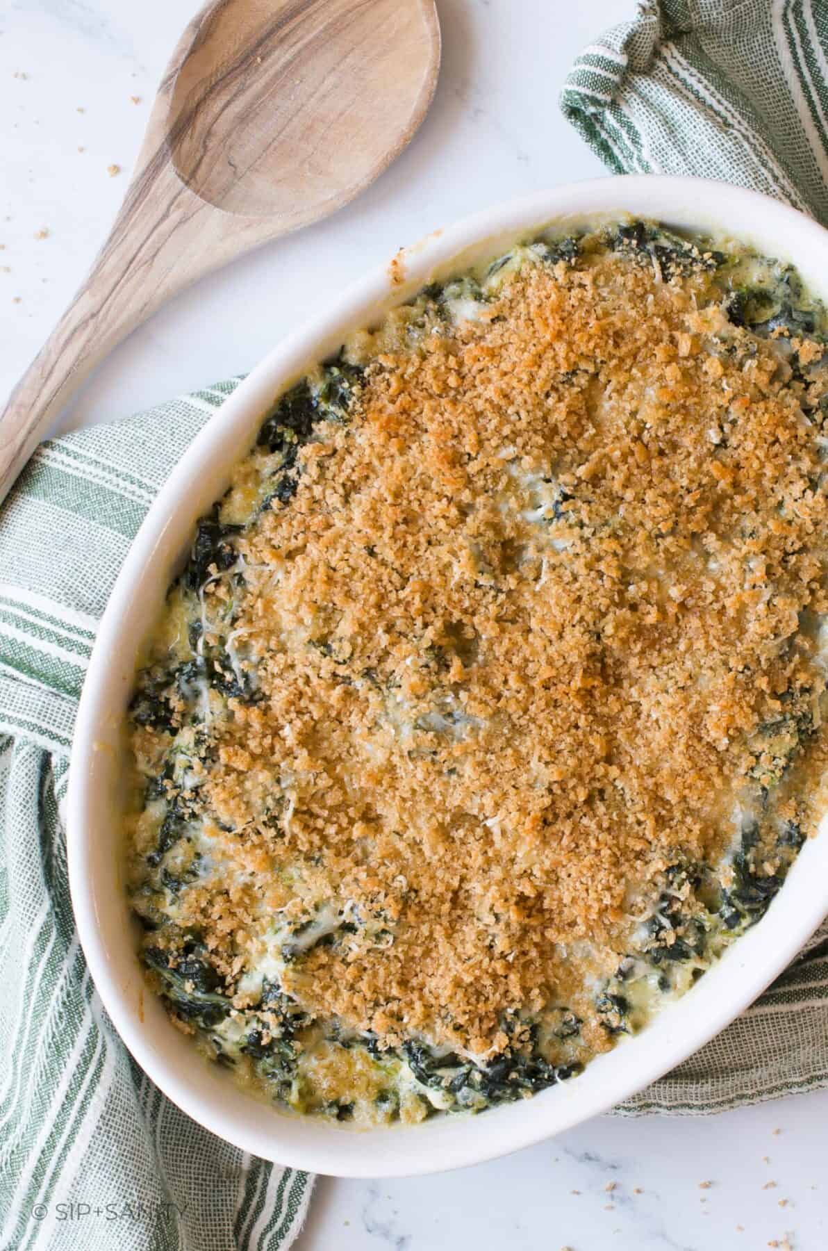 Overhead shot of asiago creamed spinach casserole on a a green striped towel next to a wooden spoon.