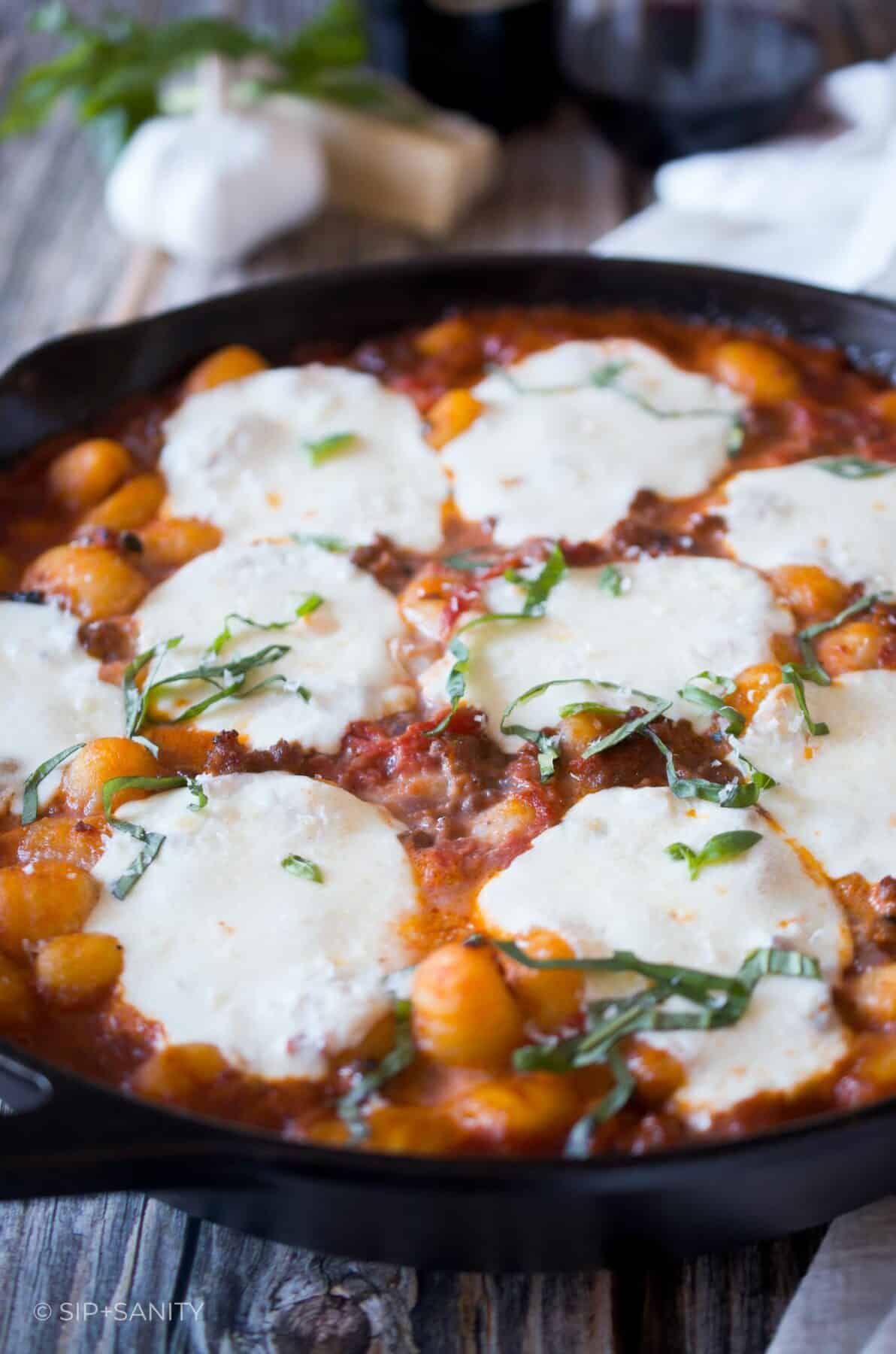 A cast iron skillet filled with gnocchi and sausage ragu with melted mozzarella and shredded basil on top.