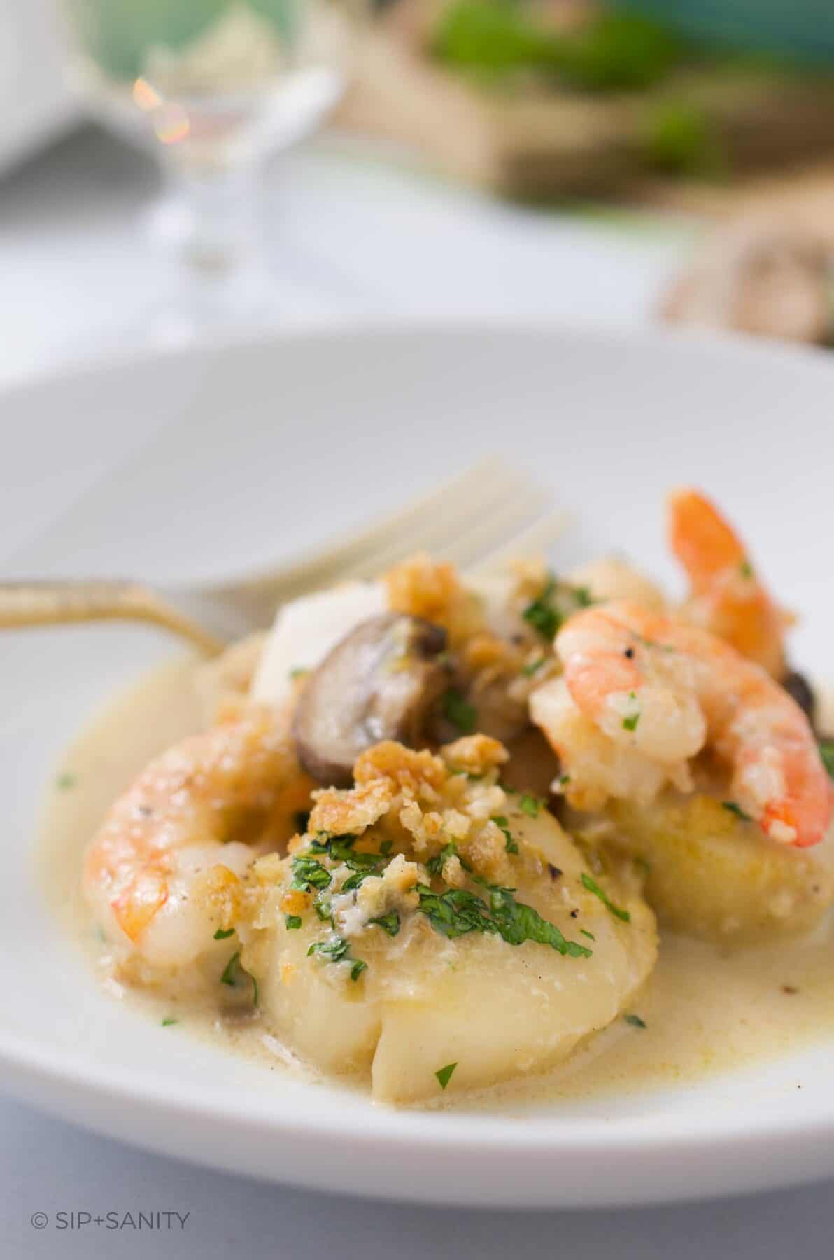 A white bowl with a scallop, shrimp, fish and mushrooms topped with breadcrumbs.