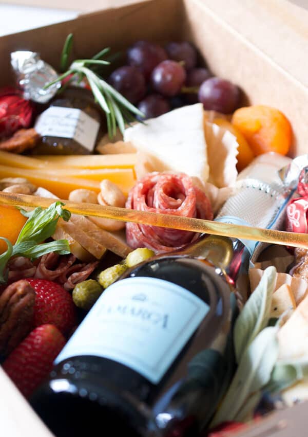 A brown paper box filled with a mini wine bottle, cheeses, salami, fruit and treats.
