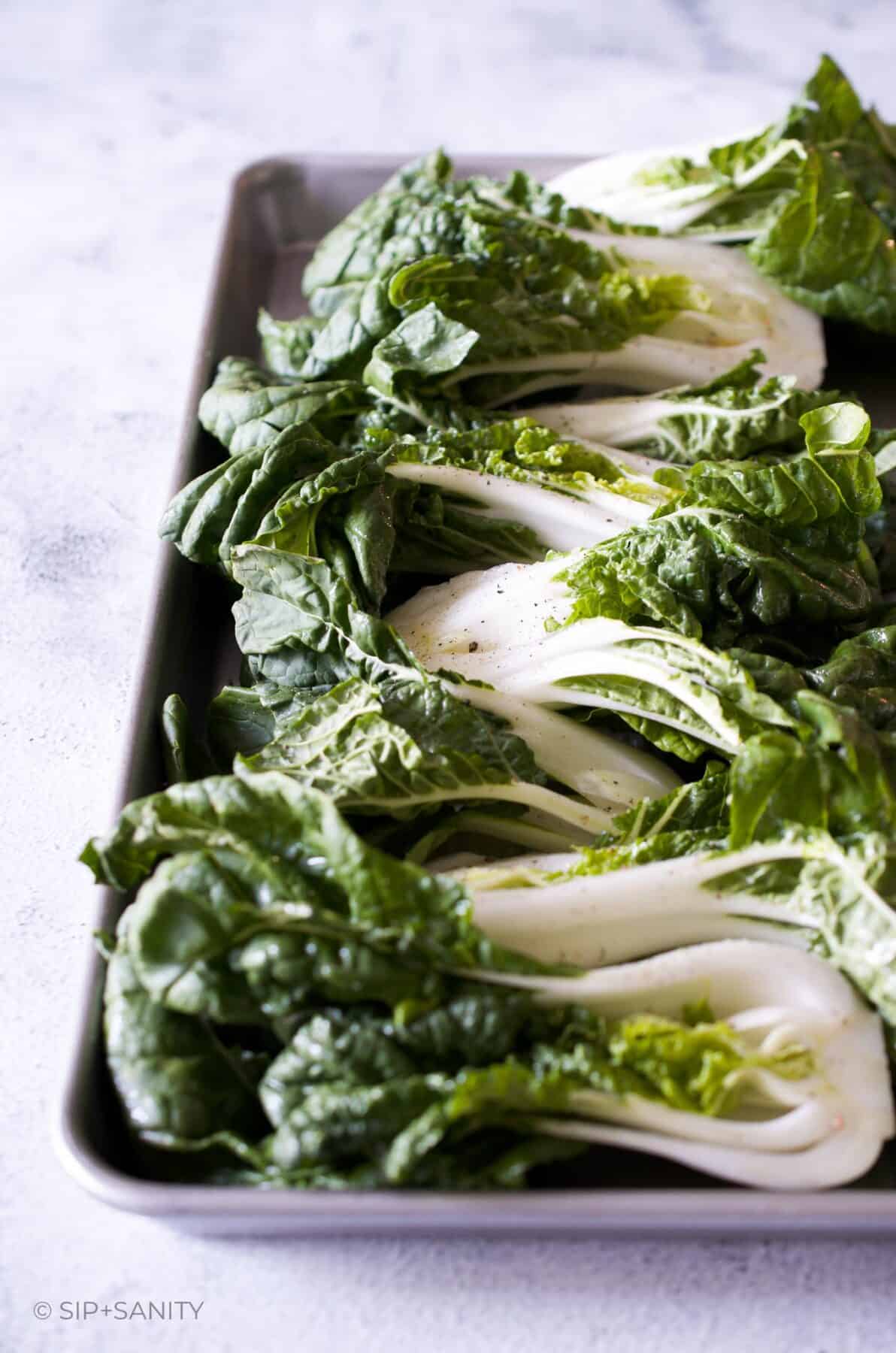 A sheet pan with an array of halved heads of baby bok choy seasoned with oil, salt and pepper.