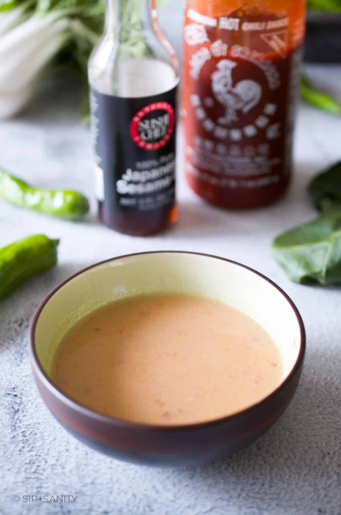 A small bowl of spicy miso finishing sauce next to bottles of sesame oil and sriracha hot sauce.