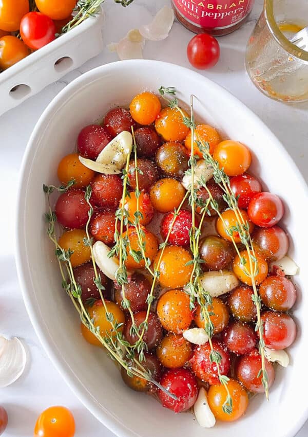 Cherry tomatoes, garlic cloves, and thyme sprigs in an oval casserole seasoned with olive oil, salt and pepper.