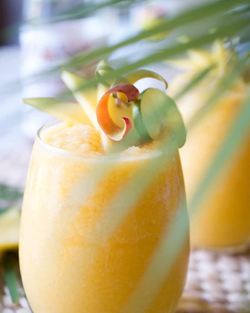 A mango colored frozen cocktail in a wine glass hiding behind a palm frond.