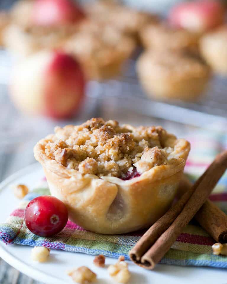 A muffin tin sized apple pie next to cinnamon sticks and a cranberry.