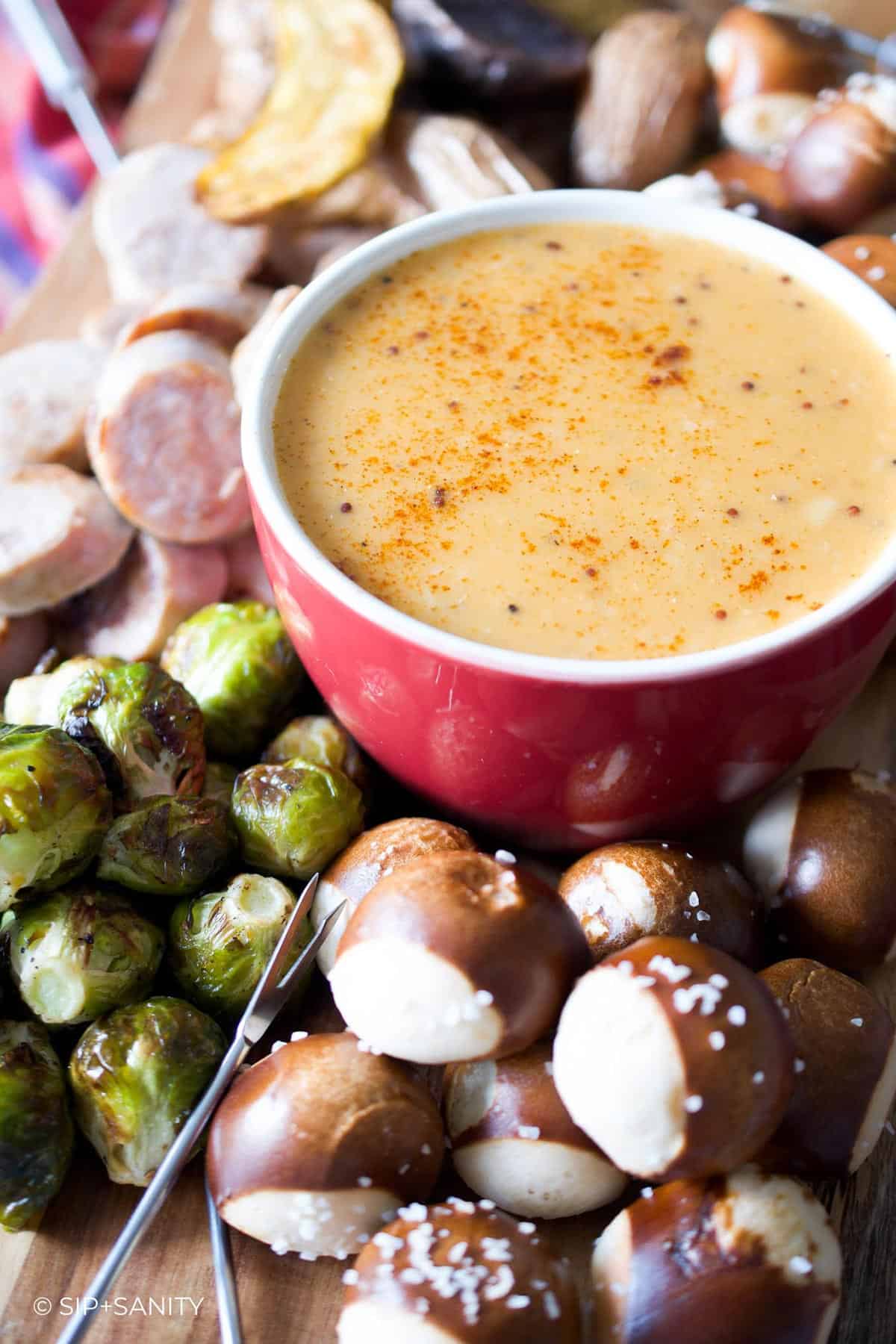 A red dish filled with beer cheese fondue surrounded by pretzels, sausage and brussels sprouts.
