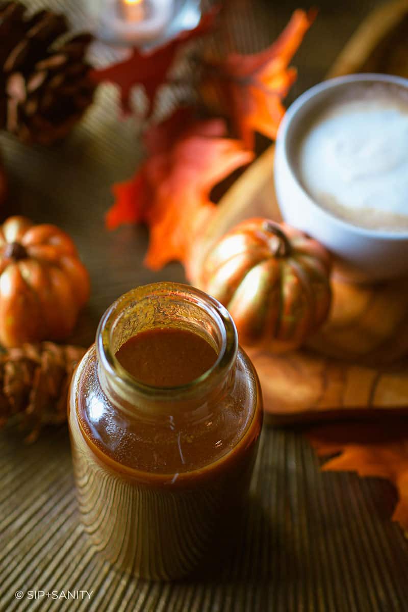 A bottle of pumpkin spice coffee syrup next to tiny pumpkins and a cup of coffee.