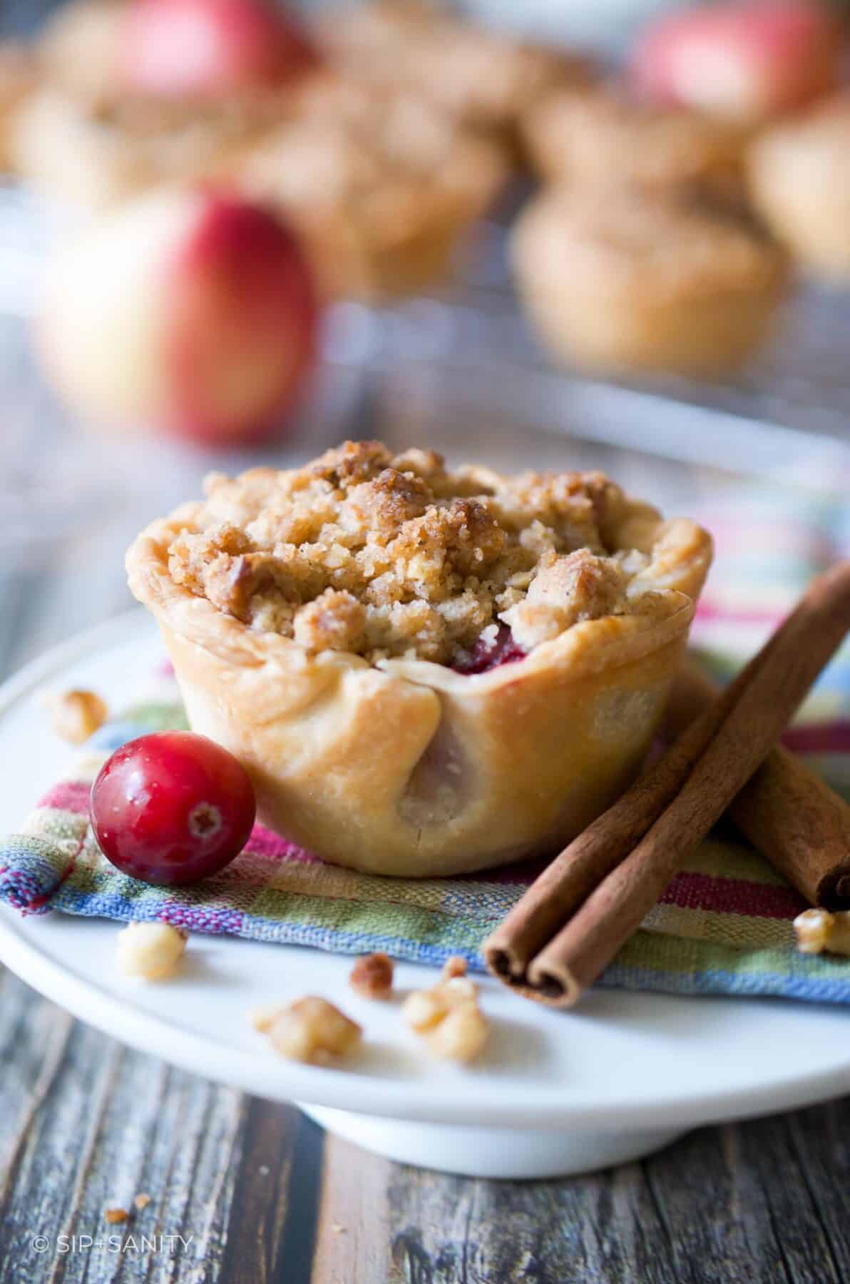 A mini apple pie on a mini cake stand with cinnamon sticks and a cranberry.