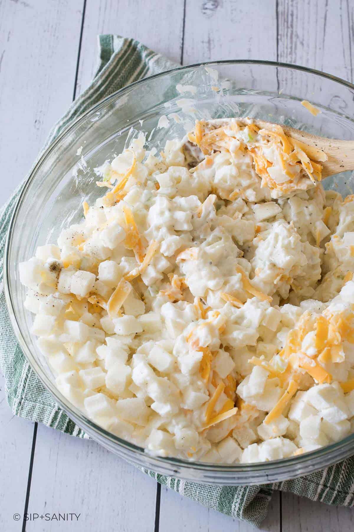 Diced potatoes, shredded cheddar and creamy sauce being mixed together in a glass bowl.
