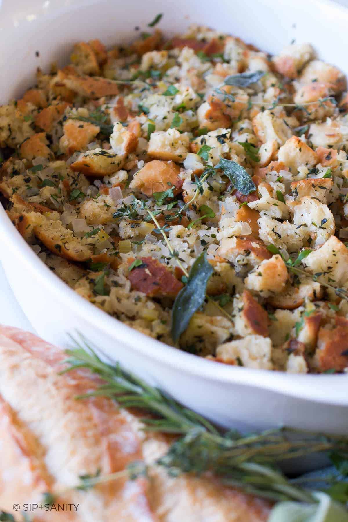A white casserole dish filled with thanksgiving stuffing next to a baguette and fresh herbs.
