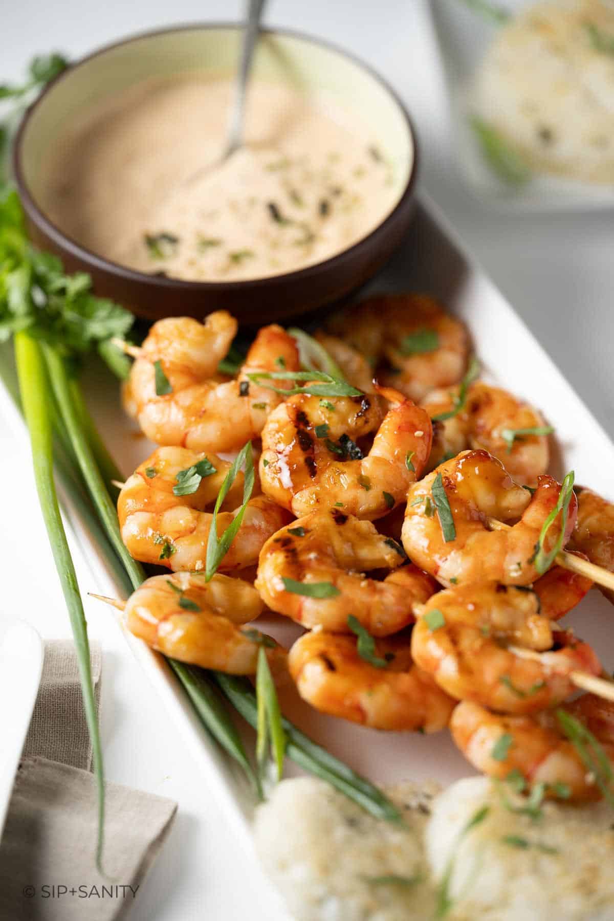 Grilled firecracker shrimp skewers next to a bowl of dipping sauce.