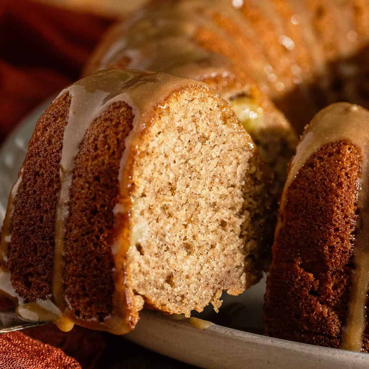 A close-up of a sliced bundt cake with a light brown interior and a shiny caramel glaze drizzling down the sides.