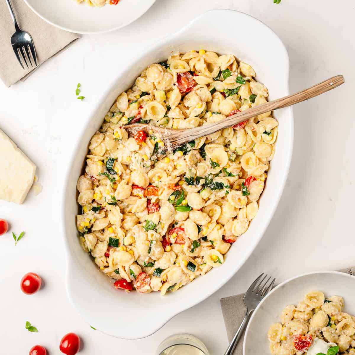 Oval white dish filled with creamy orecchiette pasta mixed with zucchini, cherry tomatoes, and corn, with a wooden spoon resting inside.