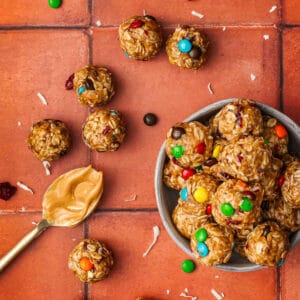 A bowl of round oatmeal energy bites with colorful candy-coated chocolates, shredded coconut, and dried cranberries sits on a terracotta tile surface.
