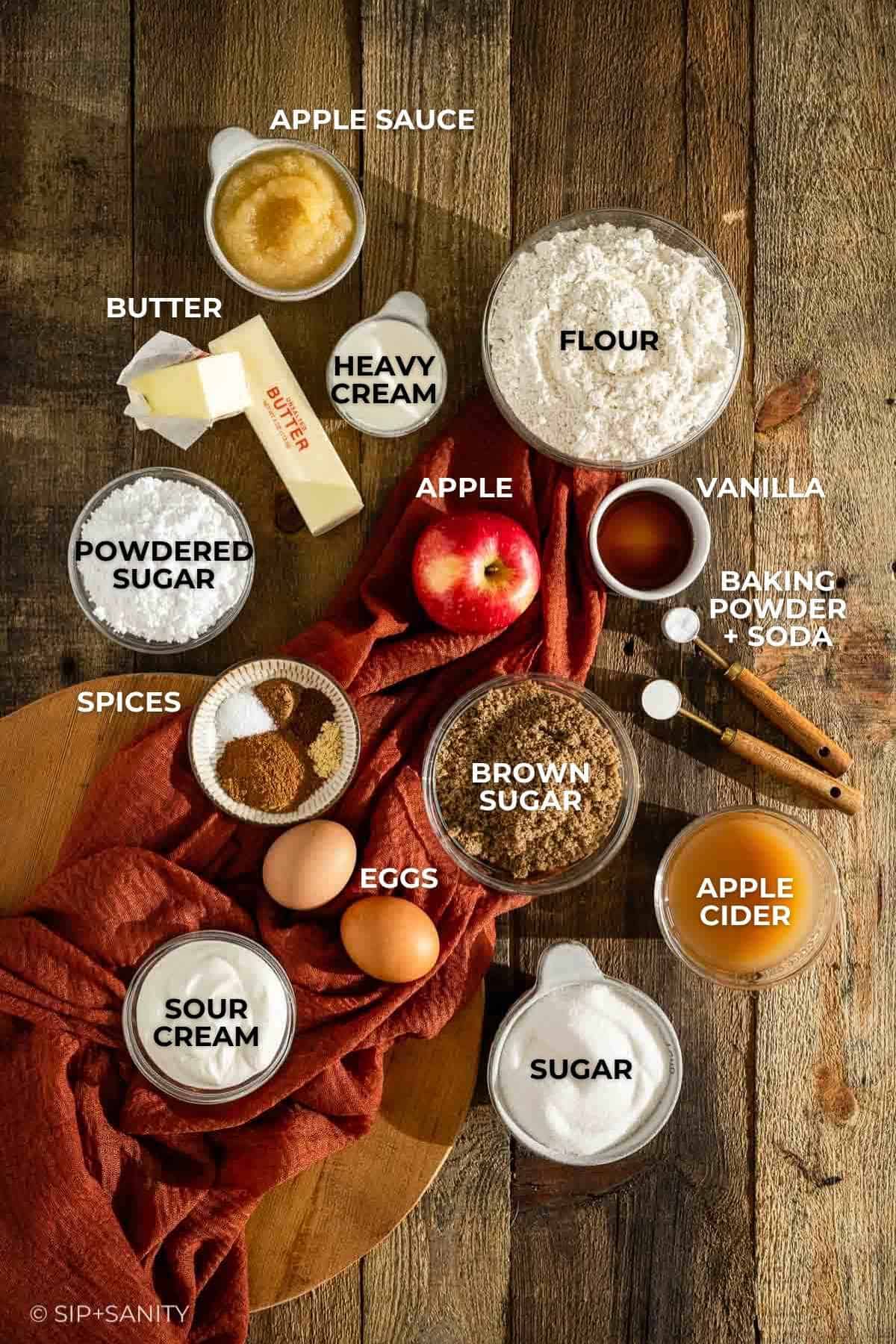 Top-down view of baking ingredients on a wooden surface, including apple sauce, flour, heavy cream, butter, powdered sugar, an apple, brown sugar, eggs, sour cream, granulated sugar, apple cider, vanilla, and baking powder and baking soda.