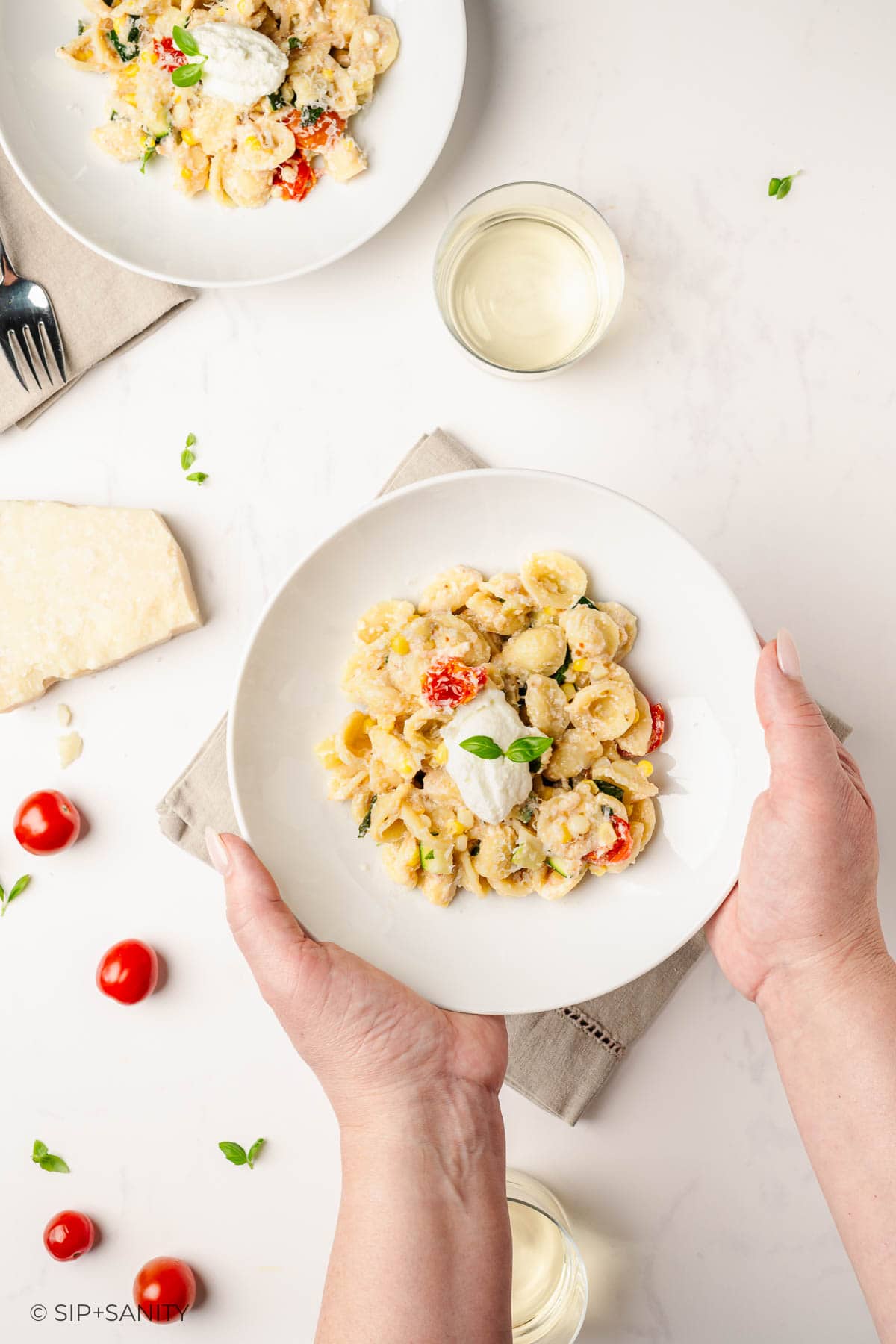 A person holds a white bowl of pasta garnished with ricotta and herbs near another bowl of pasta, a wedge of cheese, cherry tomatoes, a glass of white wine, and scattered basil leaves on a light surface.