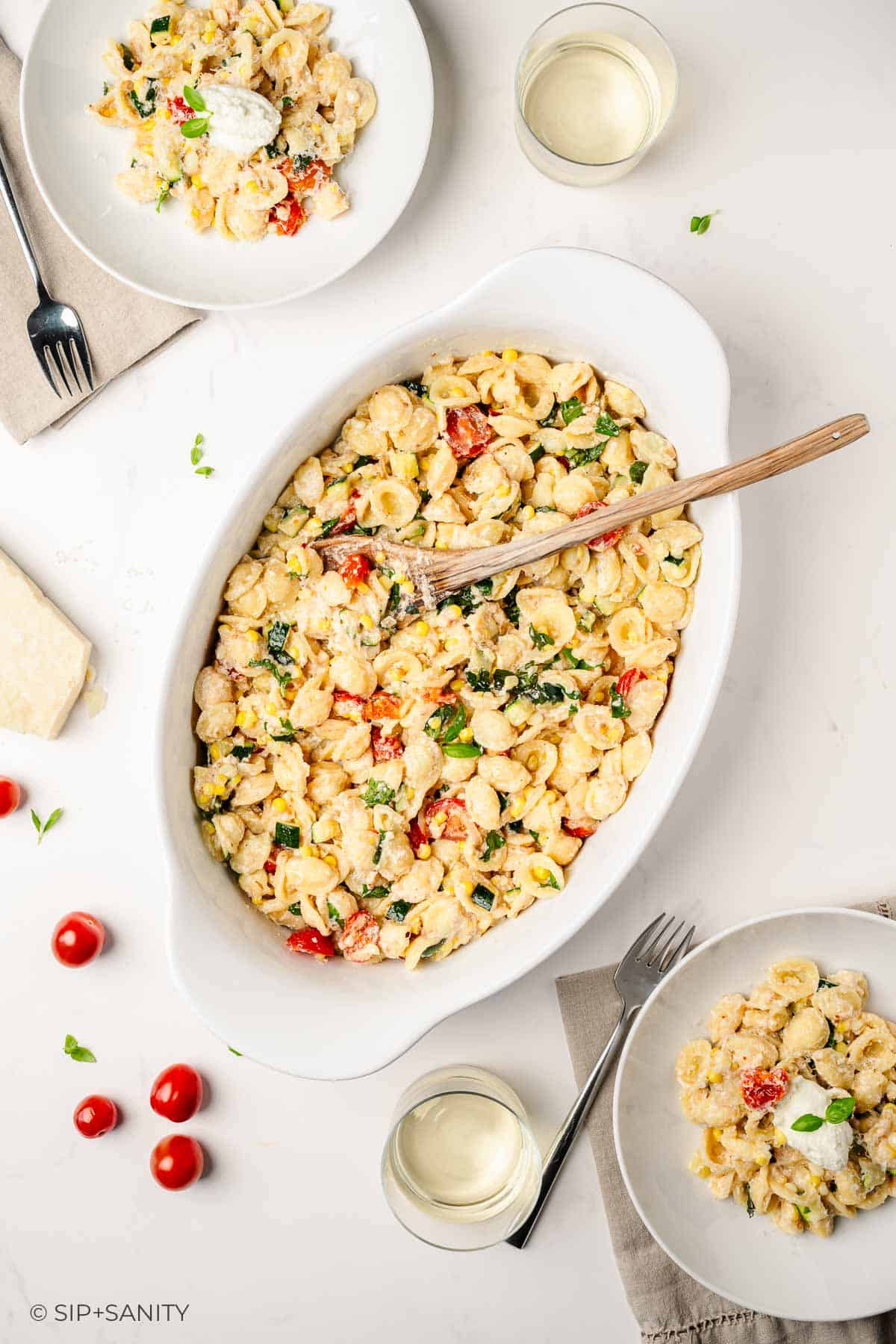Overhead view of a white oval dish filled with pasta shells, vegetables, and cheese, served with two plates of pasta, two glasses of white wine, a beige napkin, and scattered cherry tomatoes on a white table.