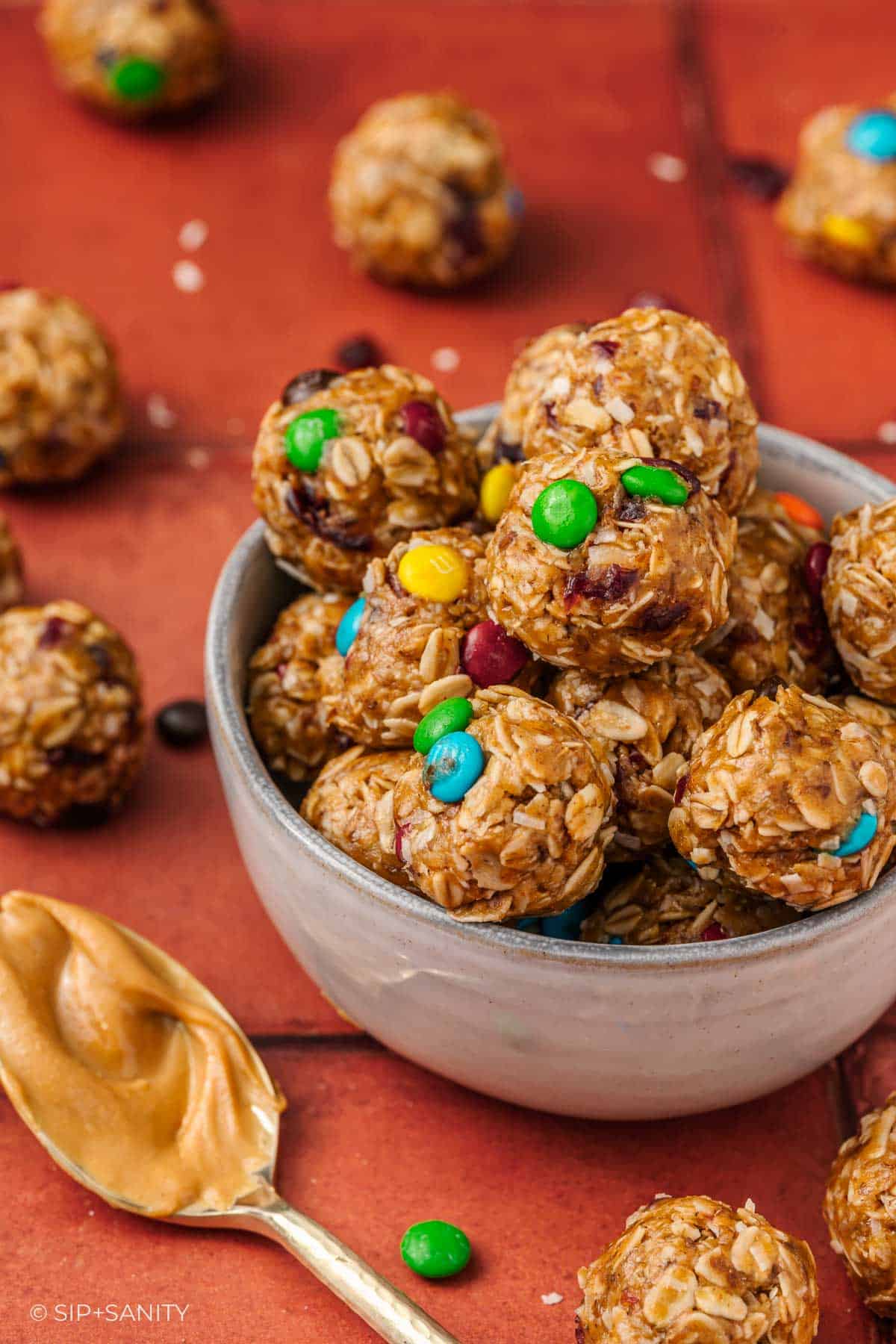 A bowl filled with oatmeal energy bites studded with colorful mix-ins, surrounded by more bites on a red tile surface.