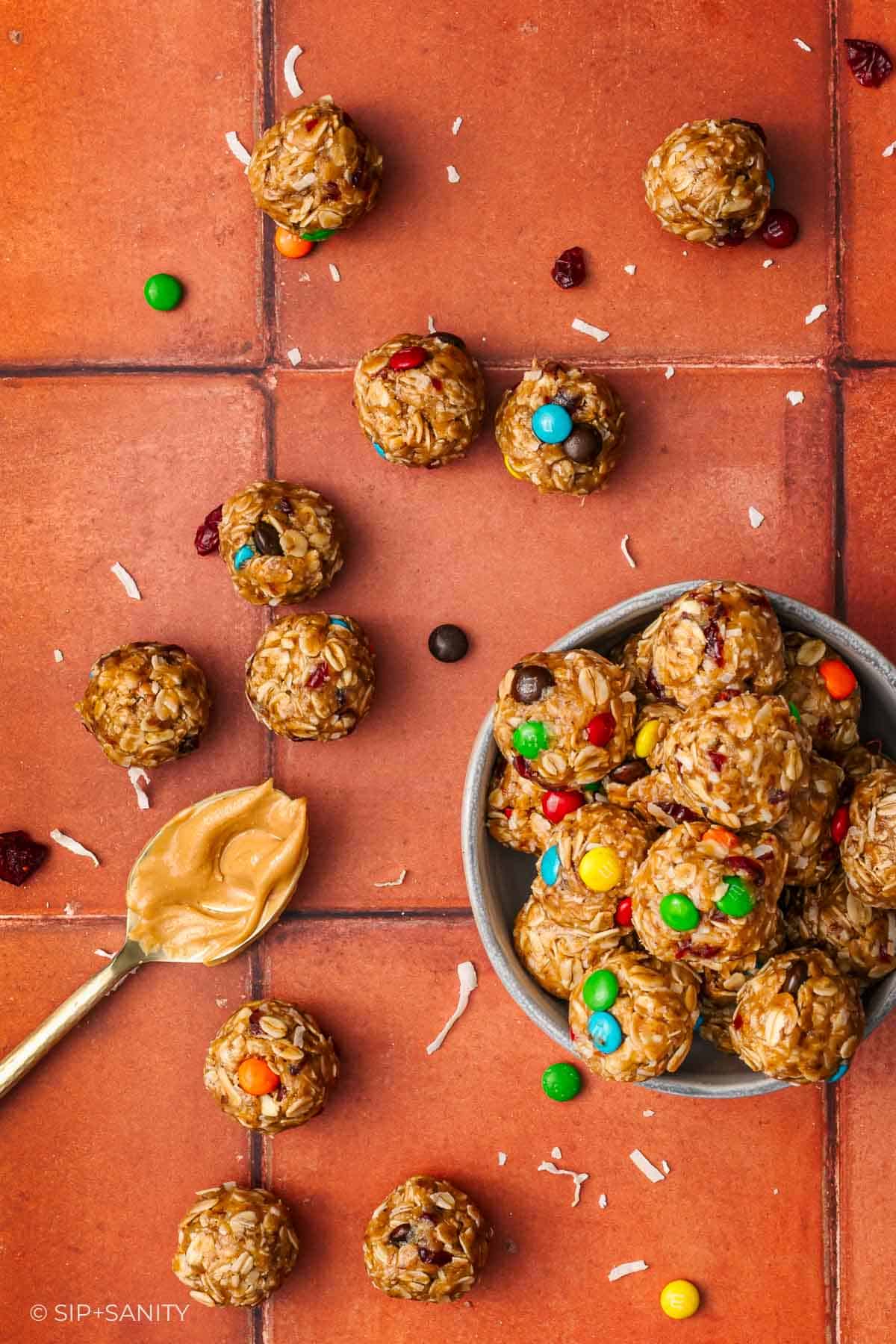Overhead view of a bowl filled with energy bites made of oats, peanut butter, and colorful candies, surrounded by more energy bites and a spoonful of peanut butter on a terra cotta tile surface.