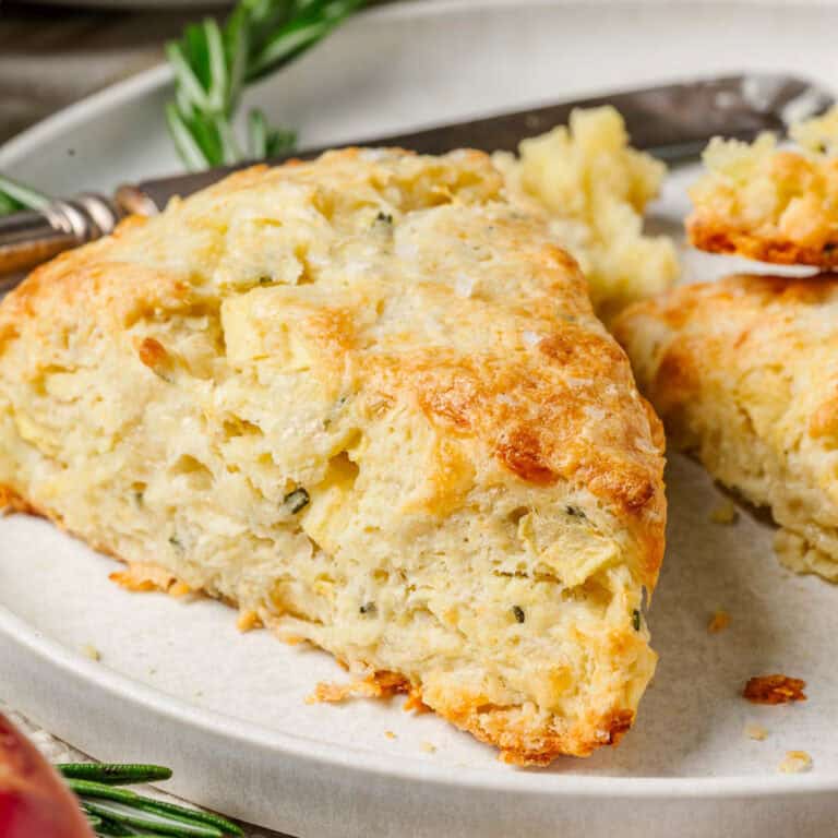 A close-up of a golden, flaky apple cheddar scone with herbs on a white plate with a knife and sprigs of fresh rosemary beside it.