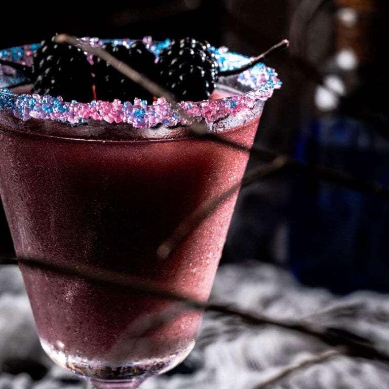 A close-up of a dark purple cocktail in a glass with a rim coated in blue and pink sugar crystals, garnished with blackberries; the background is dimly lit with a smoky, mysterious atmosphere.