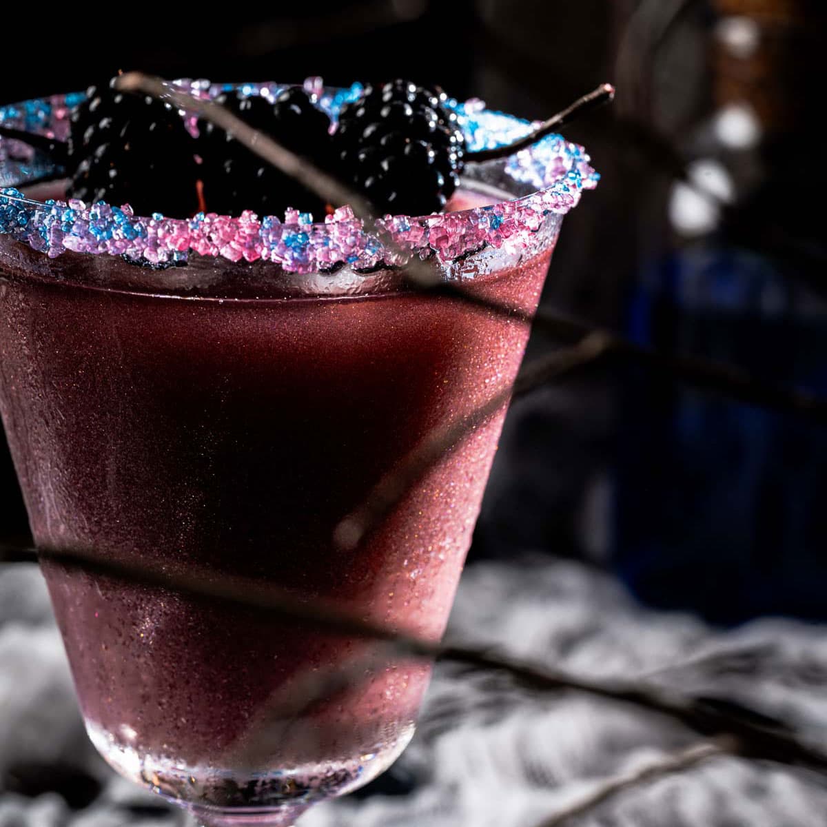 A close-up of a dark purple cocktail in a glass with a rim coated in blue and pink sugar crystals, garnished with blackberries; the background is dimly lit with a smoky, mysterious atmosphere.