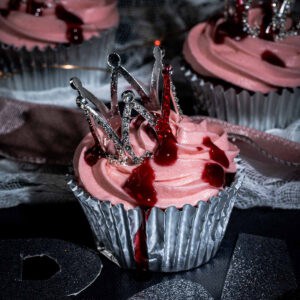 A cupcake with pink frosting in a silver wrapper, topped with a decorative silver crown and drizzled with red sauce to resemble blood, creating a dramatic and gothic appearance.
