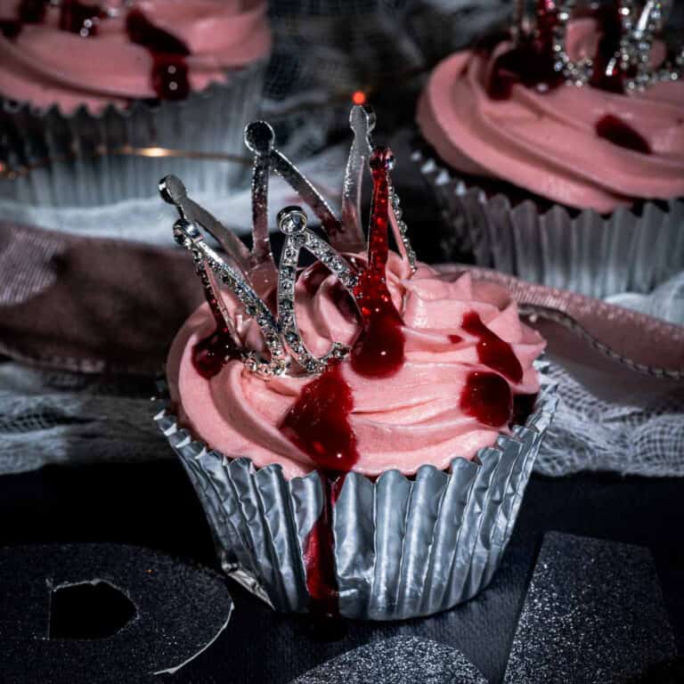 A cupcake with pink frosting in a silver wrapper, topped with a decorative silver crown and drizzled with red sauce to resemble blood, creating a dramatic and gothic appearance.