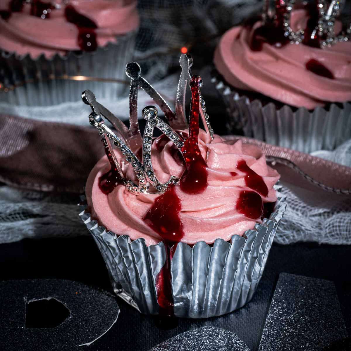 A cupcake with pink frosting in a silver wrapper, topped with a decorative silver crown and drizzled with red sauce to resemble blood, creating a dramatic and gothic appearance.