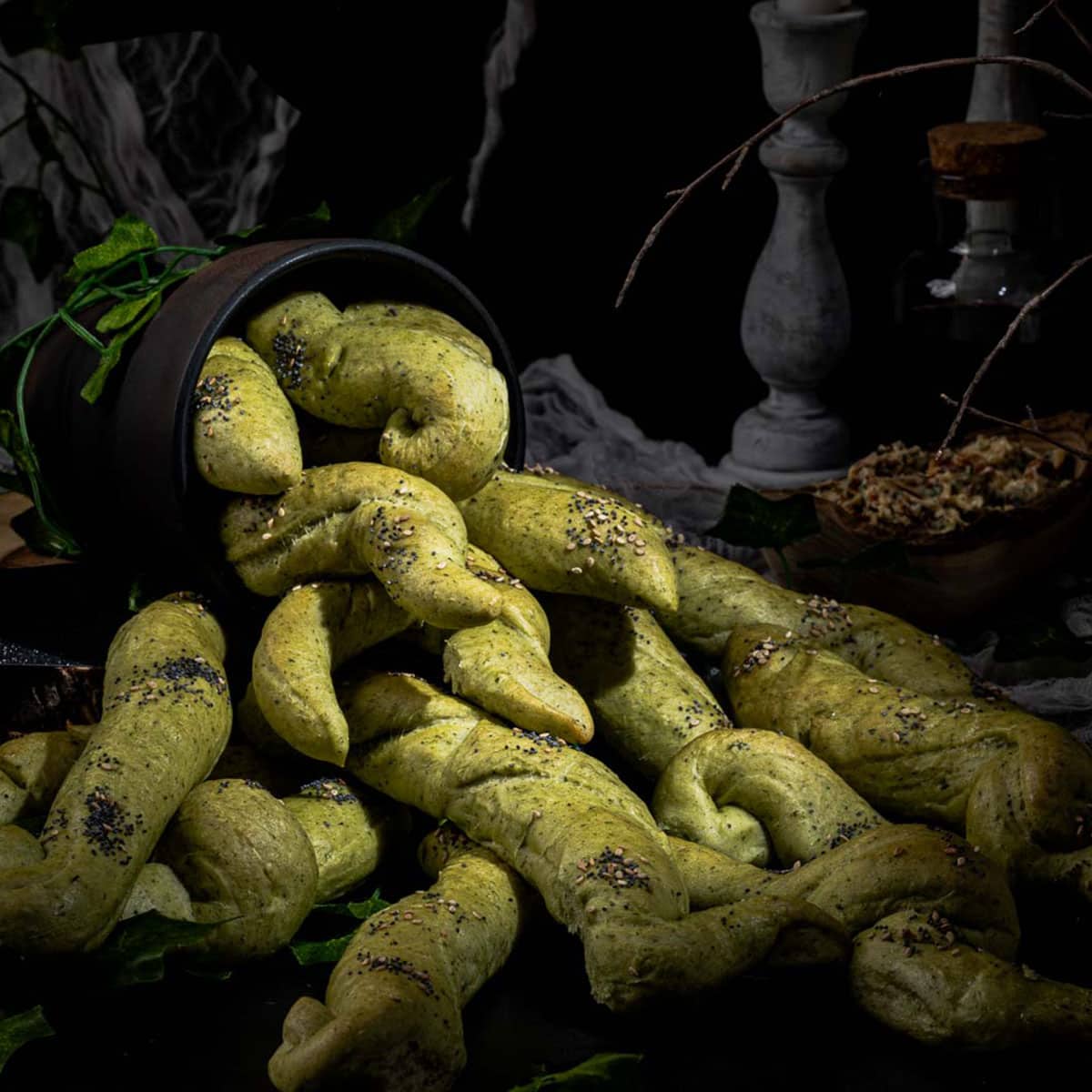 A dark ceramic pot spills out green, twisted breadsticks covered in seeds, arranged to mimic the appearance of plant tentacles on a dark surface with rustic, mysterious decor in the background.