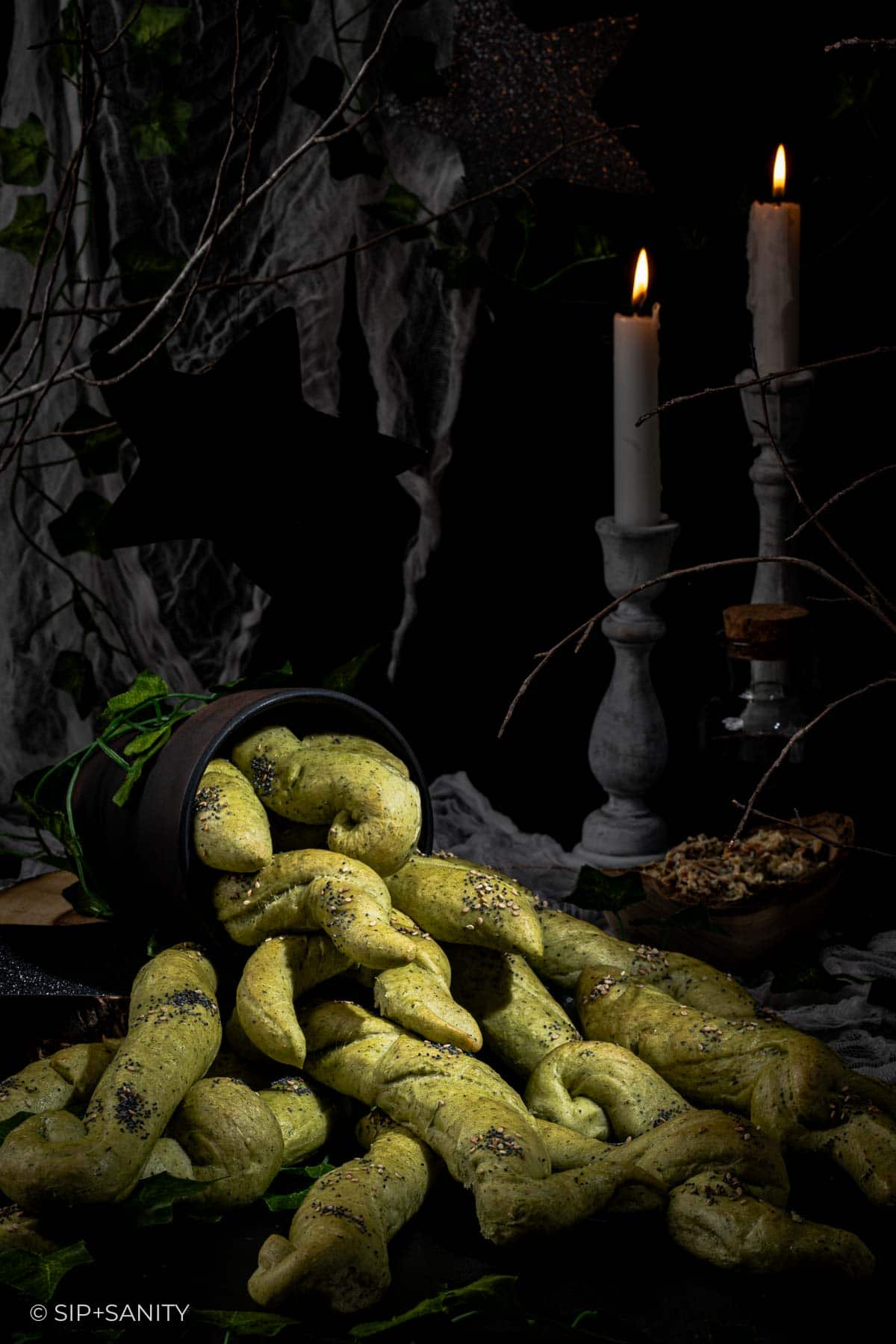 A dark, moody scene with twisted green breadsticks spilling from a black pot, surrounded by greenery, two lit candles, and rustic decor, creating a mysterious, witchy atmosphere.