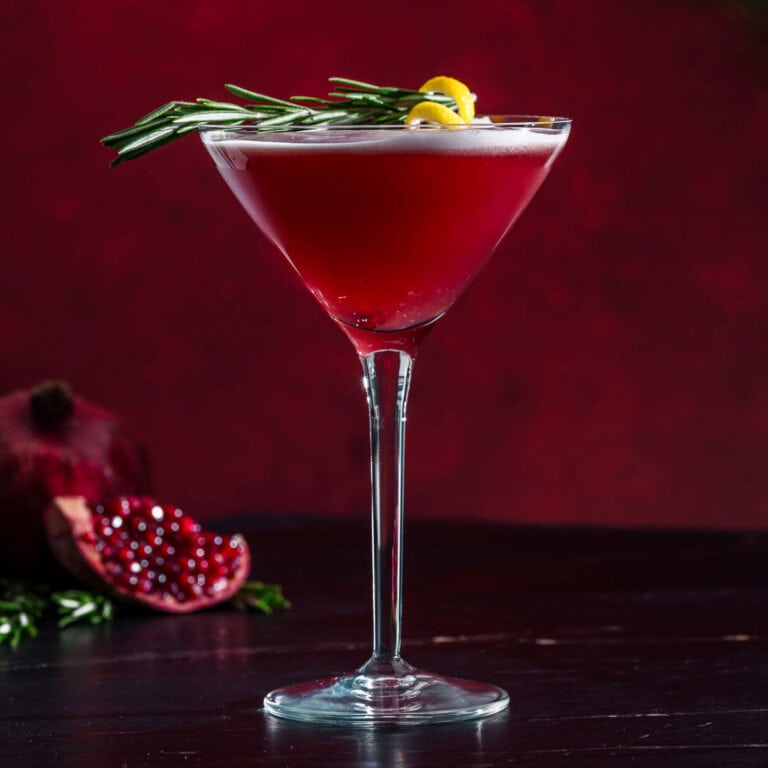 A red cocktail in a martini glass garnished with rosemary and a lemon twist sits on a dark surface, with a halved pomegranate and greenery in the background, against a deep red backdrop.