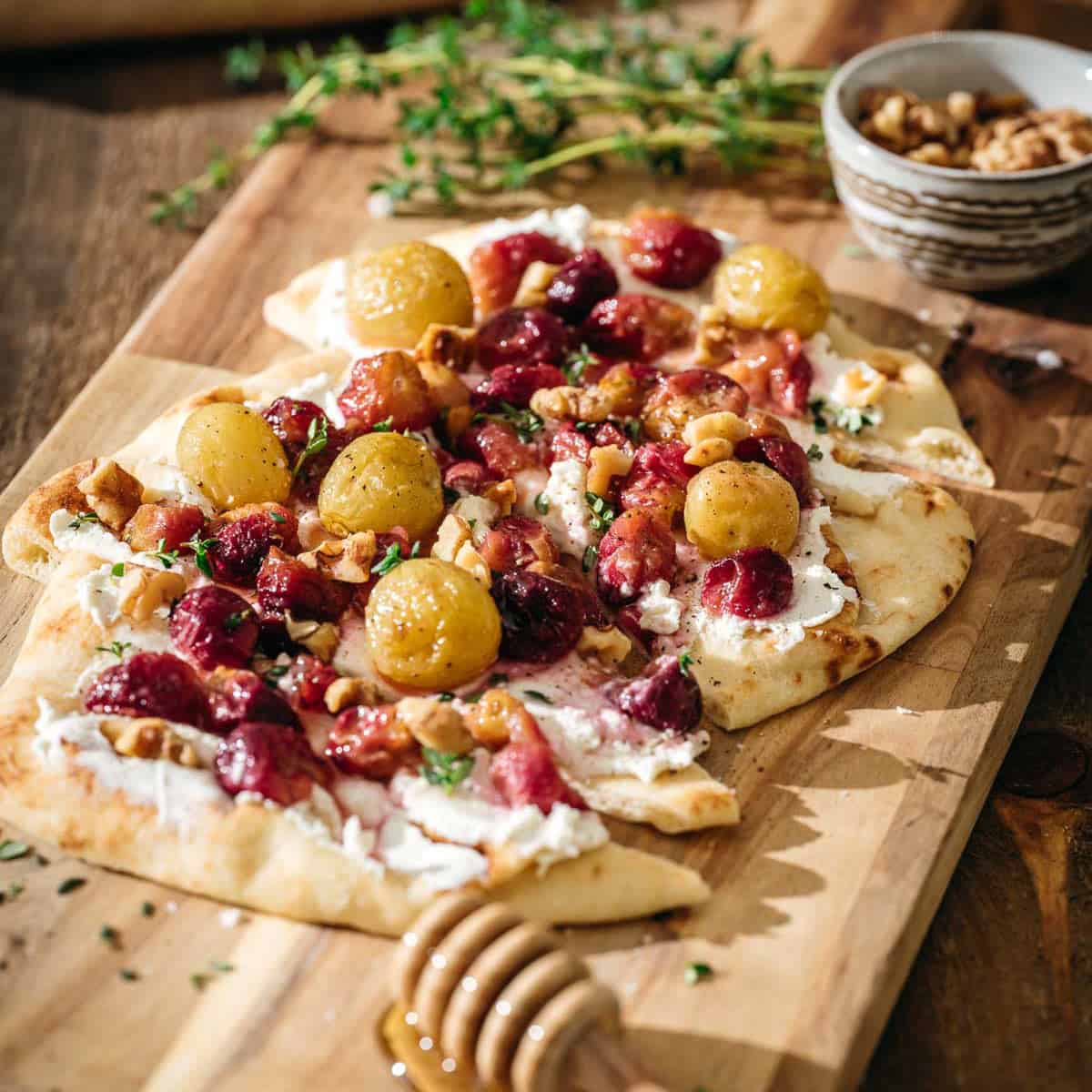 A flatbread topped with goat cheese, roasted grapes, walnuts, and herbs sits on a wooden board.