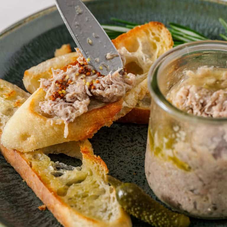 A knife spreads turkey rillette and mustard onto a toasted baguette slice.