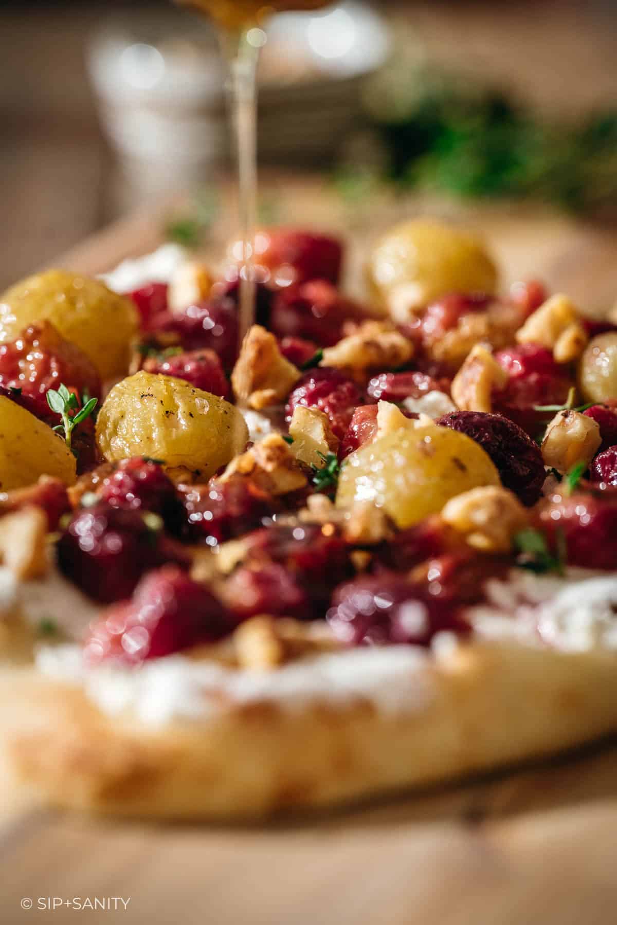 A close-up of a flatbread topped with roasted grapes, walnuts, fresh herbs, and a creamy goat cheese, with honey being drizzled over the top.